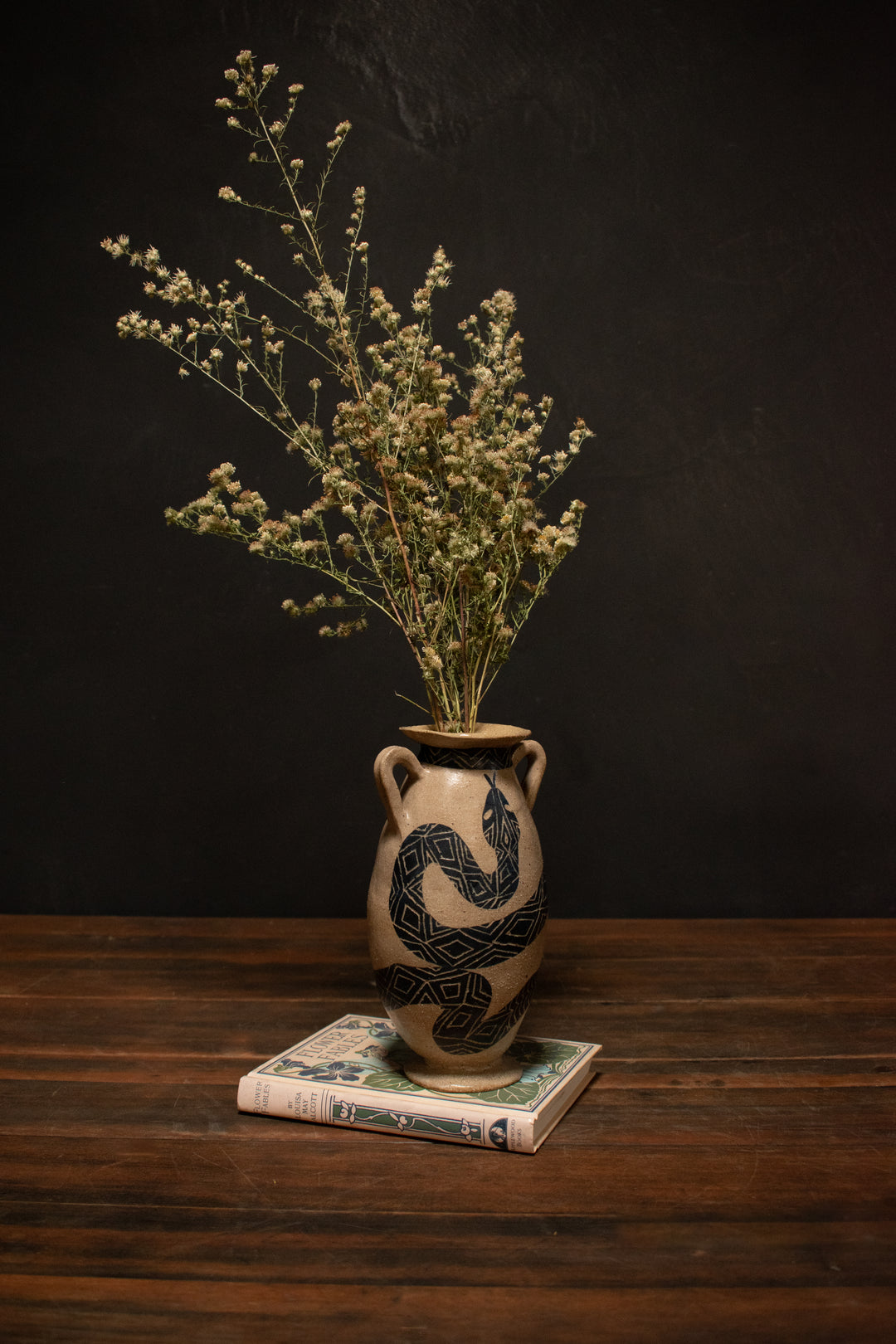 Serpent Urn With Dried Flowers sits on a vintage book in front of a black wall